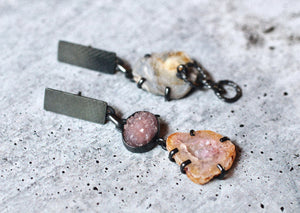 Mismatched mineral specimen earrings with amethyst crystals, agate, chalcedony rose, and oxidized silver on a textured surface.