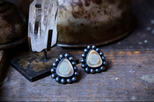 Moody studio photograph of imperial jasper eye stud earrings beside a clear quartz crystal on a metalsmithing bench.