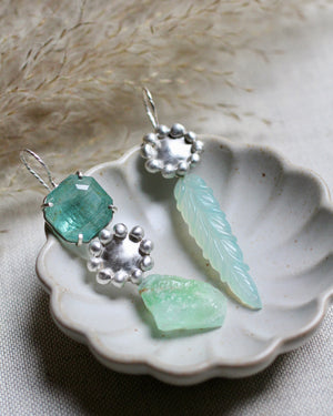 Mismatched sterling silver earrings with faceted aquamarine, raw green calcite, carved chalcedony leaf drops, and handmade satin silver baubles photographed in a jewelry dish on linen.