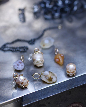 Several small skull-shaped jewelry charms and gemstone charms with gold and gemstone accents, displayed on a metallic workbench alongside beads and other jewelry.