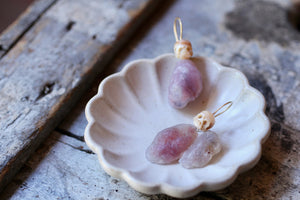 Moody close up of carved pearl skull and raw pink Gobi agate earrings resting inside a white ceramic shell dish.