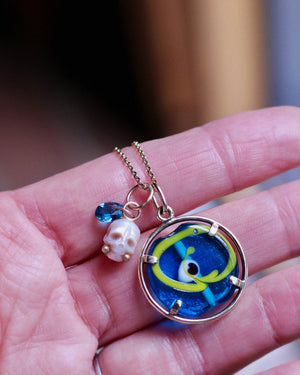 Close up of a pearl skull with gold accents and blue gemstone paired with a vintage evil eye pendant held in the hand.