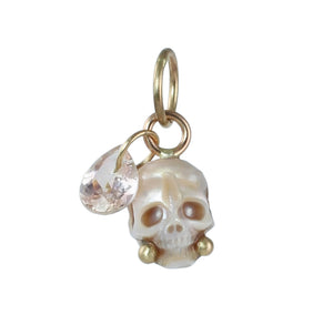 A carved pearl skull charm with gold  accents, and a dangling blush pink faceted gemstone teardrop on a plain white background.