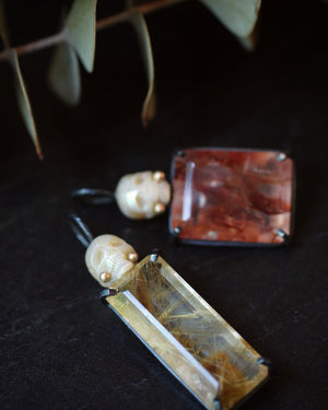 Moody close-up of two pearl Calavera pendants with red and golden rutilated quartz resting on a dark surface beneath a branch of eucalyptus.