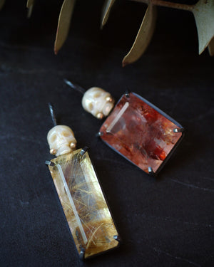 Moody close-up of two pearl Calavera pendants with red and golden rutilated quartz resting on a dark surface beneath a branch of eucalyptus.