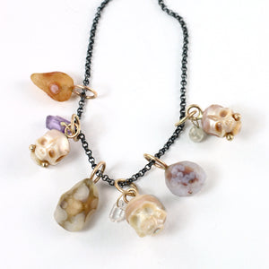 Six handcrafted charms featuring three with carved pearl skulls with gemstone and gold accents and three with colorful Gobi Desert agates shown together on an oxidized silver chain laying on a white background.