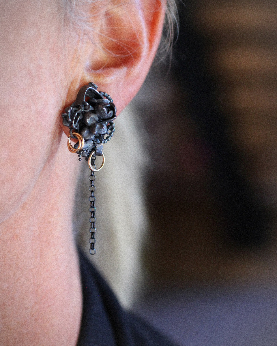 Asymmetrical Fragments earrings made from fused oxidized sterling silver with tiny flower castings, 14k gold fill rings, and dangling silver chains pictured on a clean white background.