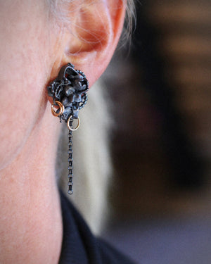 Close-up of a Fragment earring worn on the ear, showing sculptural oxidized silver, gold fill accents, and a delicate dangling chain.