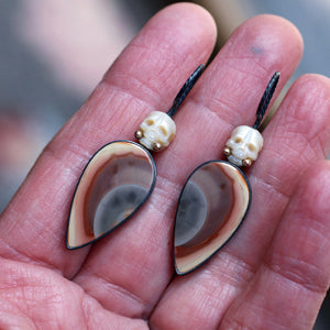 Pearl and Imperial Jasper Calavera earrings resting in a hand, with carved pearl skulls, oxidized silver ear wires, and teardrop-shaped stones in cream, rust, brown, and blue.