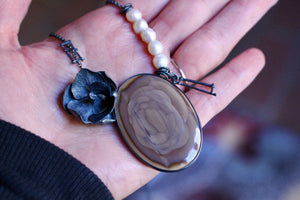 Close-up of a large mauve and brown imperial jasper with concentric rosette patterns, a dark oxidized silver orchid, irregular white pearls, and handmade ladder-like silver fragments held in a hand.