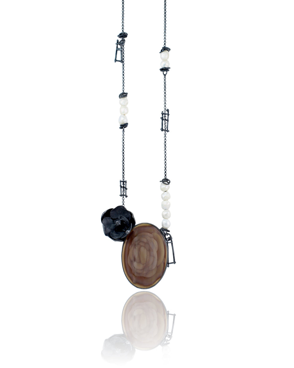 Close-up of a large mauve and brown imperial jasper with concentric rosette patterns, a dark oxidized silver orchid, irregular white pearls, and handmade ladder-like silver fragments. Photographed floating on a white background.