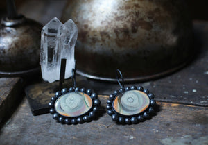 Imperial jasper eye earrings displayed on a metalsmithing bench beside a clear quartz crystal and aged studio tools.