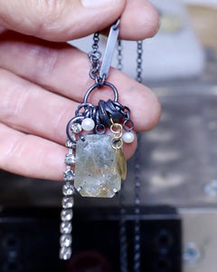Video close-up of a handmade necklace held in hand, featuring a faceted green sunstone with coppery-gold shimmer, pearls, oxidized sterling silver fragments and dangling crystal chain. Shows movement and sparkle of the sunstone.