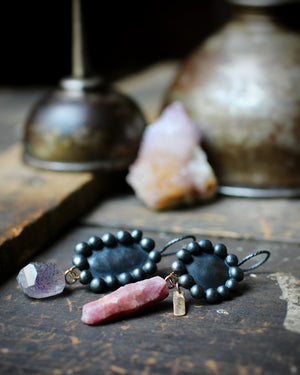 Raw ruby crystal and melody stone earrings in oxidized sterling silver with recycled 14k gold accents, photographed on a dark studio surface with antique items and a stone in the background.