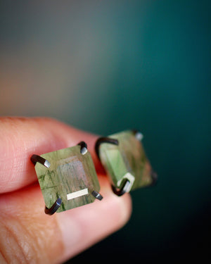 Close-up of faceted green rutilated quartz studs held between fingers, showing their natural threadlike inclusions.