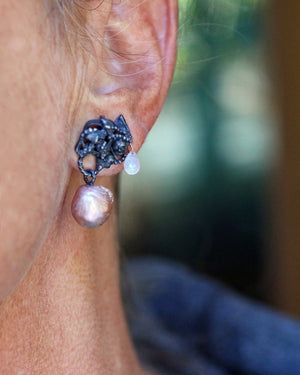 Close-up of mismatched fused sterling silver earrings worn on the ear, showing a hidden skull, luminous pink pearl, faceted moonstone drop, and 14k gold detail.