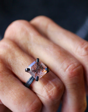 Handmade modernist sunstone ring with warm orange and copper inclusions in a dark oxidized sterling silver setting, shown on a hand.