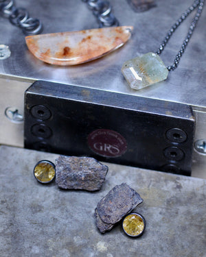 A close-up of jewelry pieces arranged on a metal workbench, including a crescent-shaped peach stone pendant on a chain, a pale green rectangular stone pendant on a black chain, and two small round gold-toned bezel-set stones set in rough gray rock.
